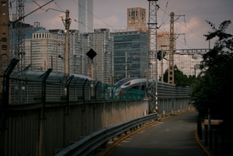 A sleek, modern train travels along a track surrounded by urban high-rise buildings. Various electrical wires and poles are visible overhead, indicative of a bustling metropolitan setting. The pathway beside the tracks is lined with protective barriers.