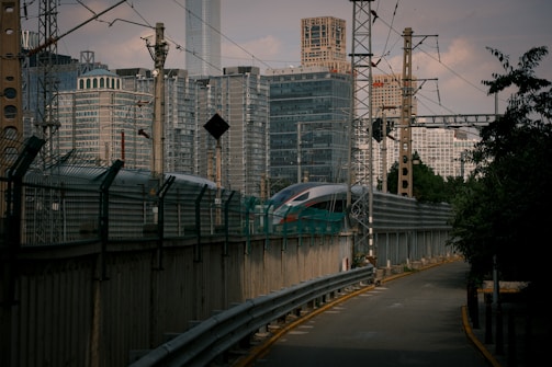 A sleek, modern train travels along a track surrounded by urban high-rise buildings. Various electrical wires and poles are visible overhead, indicative of a bustling metropolitan setting. The pathway beside the tracks is lined with protective barriers.