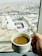 A hand holding a white ceramic cup filled with coffee, with a large cityscape visible through a window in the background. The city features a vast complex with a white tent-like structure and multiple minarets, surrounded by hills in the distance.