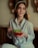 A woman with long brown hair and hoop earrings is smiling gently. She is wearing a light blue shirt over a white top, and holding a red bowl filled with green grapes.