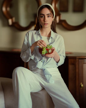 A serene woman in a light, flowing outfit enjoying a sunny morning with a bowl of fresh, colorful fruits on a minimalist wooden table.