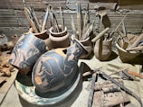 A variety of artist tools and pottery on a wooden surface. There are several broken ceramics with intricate black and orange designs featuring fish motifs. Surrounding the pottery are multiple brushes and tools stored in rustic clay containers. The background shows a textured brick wall, adding to the rustic and artistic atmosphere.