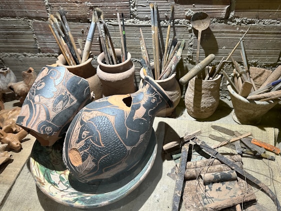 A variety of artist tools and pottery on a wooden surface. There are several broken ceramics with intricate black and orange designs featuring fish motifs. Surrounding the pottery are multiple brushes and tools stored in rustic clay containers. The background shows a textured brick wall, adding to the rustic and artistic atmosphere.