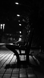 A monochrome photograph of an empty bench lined along a pavement. Streetlights illuminate the pathway, casting shadows and creating a moody atmosphere. The background fades into darkness, with trees and soft light visible.