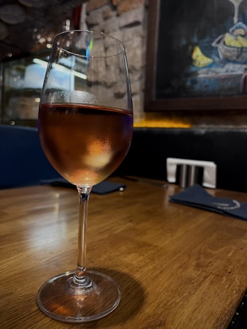 A glass of amber-colored wine rests on a wooden table. The background features a rustic wall with exposed bricks and dim lighting, creating a cozy ambiance. A framed artwork with blue and yellow hues hangs on the wall.