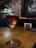 A glass of amber-colored wine rests on a wooden table. The background features a rustic wall with exposed bricks and dim lighting, creating a cozy ambiance. A framed artwork with blue and yellow hues hangs on the wall.