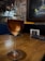 A glass of amber-colored wine rests on a wooden table. The background features a rustic wall with exposed bricks and dim lighting, creating a cozy ambiance. A framed artwork with blue and yellow hues hangs on the wall.