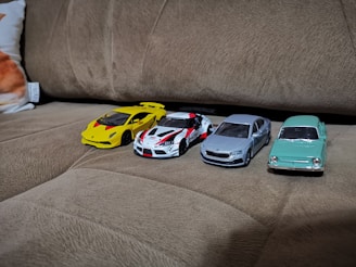 Four toy cars are arranged side by side on a brown couch. The cars include a yellow sports car, a white and red racing car, a metallic silver sedan, and a light green vintage car. The texture of the couch is soft and textured with visible stitching lines.