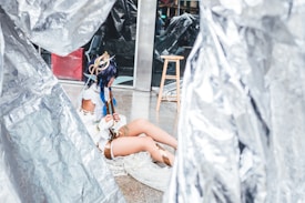 A person wearing a detailed fantasy costume sits on the ground surrounded by metallic-looking large sheets. The costume features accessories like a headpiece with ribbons and feathers and clothing with white and brown elements. Nearby, there is a wooden stool and reflective surfaces.