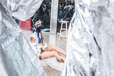 A person wearing a detailed fantasy costume sits on the ground surrounded by metallic-looking large sheets. The costume features accessories like a headpiece with ribbons and feathers and clothing with white and brown elements. Nearby, there is a wooden stool and reflective surfaces.