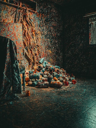 A dimly lit room with walls covered in splattered paint in various colors. There is a messy arrangement of paint cans and bottles piled up on the floor. Streaks of paint cover the walls, creating a chaotic and artistic environment. The lighting casts shadows that enhance the cluttered and creative atmosphere.