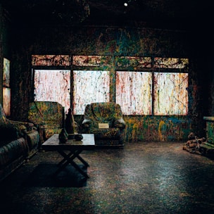 A room with furniture covered in multicolored paint splatters, including two armchairs and a couch. There is a coffee table with vases and a small sign. The windows are also splattered with colorful paint, creating a chaotic and artistic effect.