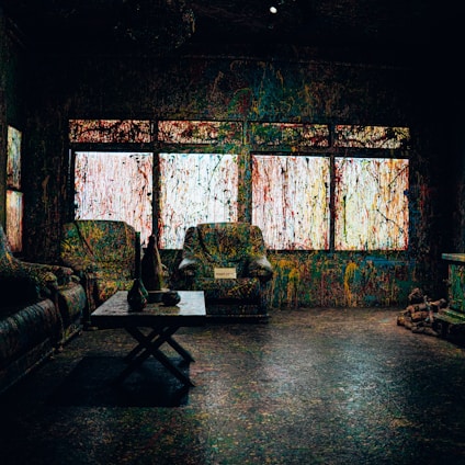 A room with furniture covered in multicolored paint splatters, including two armchairs and a couch. There is a coffee table with vases and a small sign. The windows are also splattered with colorful paint, creating a chaotic and artistic effect.