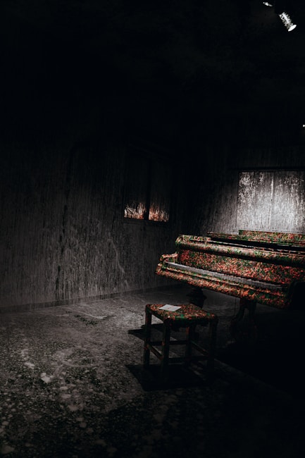 A dimly lit room features a piano and a stool, both covered in a vibrant floral pattern. The walls and floor have a textured, weathered appearance, contributing to a surreal and isolated atmosphere. A small square window allows a subtle glow to filter through, adding to the mysterious ambiance.
