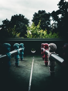 A foosball table viewed from the perspective of the players, with blue and red figurines lined up on opposing rods. The table is outdoors with blurred trees and greenery in the background, suggesting it might be in a garden or park. The lighting is soft and overcast, creating a somewhat moody atmosphere.