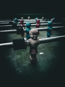 A close-up of a foosball table with figurines attached to metal rods. The figurines are painted in worn colors, some in shades of red and others in blue-green. The background is dark, creating a dramatic contrast with the figures.