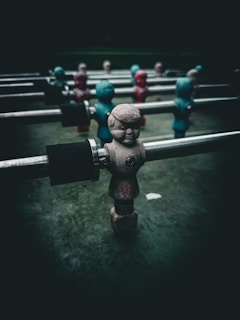 A close-up of a foosball table with figurines attached to metal rods. The figurines are painted in worn colors, some in shades of red and others in blue-green. The background is dark, creating a dramatic contrast with the figures.