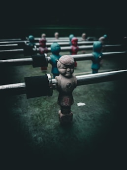 A close-up of a foosball table with figurines attached to metal rods. The figurines are painted in worn colors, some in shades of red and others in blue-green. The background is dark, creating a dramatic contrast with the figures.