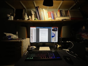 A dimly lit desk setup featuring a computer monitor displaying documents. The desk is organized with bottles, a keyboard with illuminated keys, and various stationery. Above the desk is a shelf filled with books and small decorative items.