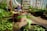 A vibrant greenhouse filled with various potted plants and greenery. In the foreground, a young plant is growing in a pot, and a child in warm clothing is blurred in the background, suggesting activity or care in the garden. The space is well-lit, with pots of different sizes and plants arranged neatly.