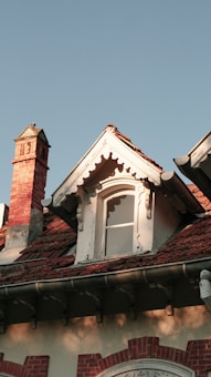A charming rooftop scene featuring a gabled window with intricate woodwork and a small chimney constructed from red bricks. The roof is covered with weathered reddish-brown tiles that give a rustic feel. The architecture appears vintage, possibly from the Victorian era, set against a clear blue sky.