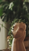 A clay sculpture of a human head acts as a planter, with a trailing succulent cascading over its top, resembling hair. The background is filled with lush, blurred greenery, giving an impression of a natural indoor setting.