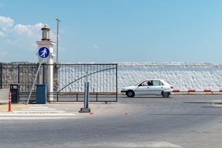 A white car is waiting at a barrier near a pedestrian crossing sign. The area is fenced with black metal, and there is a lamp post nearby. A person is visible inside the vehicle. The street is wide, and the background features a tall white wall under a blue sky with some clouds.
