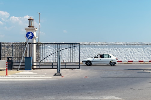 A white car is waiting at a barrier near a pedestrian crossing sign. The area is fenced with black metal, and there is a lamp post nearby. A person is visible inside the vehicle. The street is wide, and the background features a tall white wall under a blue sky with some clouds.