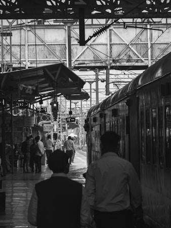 A bustling train station platform with numerous individuals walking and standing around. A train is parked on the right side of the image, and various overhead structures and signage are visible. The image is in black and white, adding a classic, timeless feel.