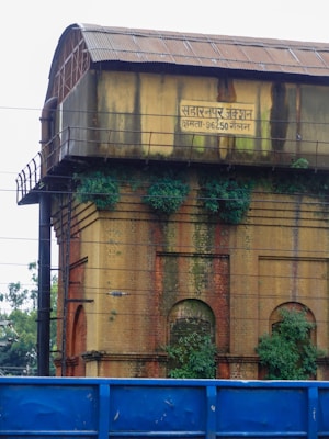 An old brick structure with a corrugated metal roof and a sign in Hindi. The building shows signs of weathering and is partially covered in green vegetation. A bright blue barrier or fence is visible in the foreground.