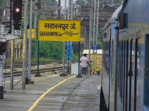 A train is stationed at a railway platform with a yellow sign displaying 'Saharanpur Jn.' in multiple languages. There's a railway signal and tracks on the left, and a person is visible on the platform wearing a pink shirt.
