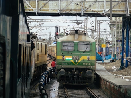 A green locomotive is prominently featured at a railway station, surrounded by tracks, overhead wires, and a pedestrian bridge. Another train is visible on the left. Rail infrastructure, signage, and basic station facilities are visible.