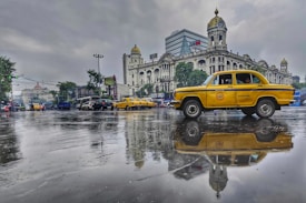 A vintage yellow taxi is prominently reflected on a wet road, with a backdrop of a grand colonial-style building adorned with a clock tower. The scene features overcast skies with a moody atmosphere, and other cars and buildings can be seen in the background.