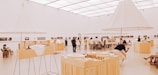 An art gallery with a modern, minimalist design features several people admiring exhibits. The room is spacious and well-lit, with large, white, conical light fixtures and wooden display tables showcasing architectural models and various artworks.