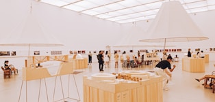 An art gallery with a modern, minimalist design features several people admiring exhibits. The room is spacious and well-lit, with large, white, conical light fixtures and wooden display tables showcasing architectural models and various artworks.