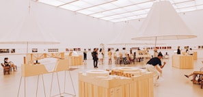 An art gallery with a modern, minimalist design features several people admiring exhibits. The room is spacious and well-lit, with large, white, conical light fixtures and wooden display tables showcasing architectural models and various artworks.