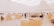 An art gallery with a modern, minimalist design features several people admiring exhibits. The room is spacious and well-lit, with large, white, conical light fixtures and wooden display tables showcasing architectural models and various artworks.