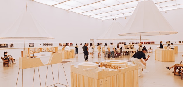 An art gallery with a modern, minimalist design features several people admiring exhibits. The room is spacious and well-lit, with large, white, conical light fixtures and wooden display tables showcasing architectural models and various artworks.