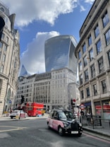 A bustling urban scene featuring modern skyscrapers, historic buildings, a red double-decker bus, and a black London taxi with pink advertisement. The scene captures the essence of a busy street surrounded by architectural landmarks under a blue sky with scattered clouds.