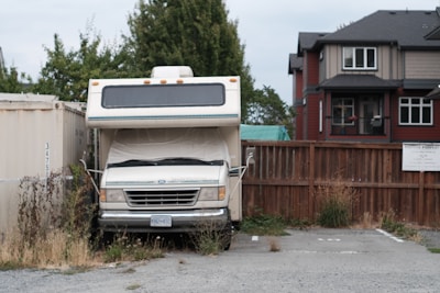 An older model RV is parked in a small, overgrown parking space adjacent to a wooden fence. The RV is flanked by a shipping container and multiple storage units. Weeds and grass are growing around the base of the vehicle. In the background, there is a modern two-story house with dark roofing.