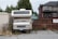 An older model RV is parked in a small, overgrown parking space adjacent to a wooden fence. The RV is flanked by a shipping container and multiple storage units. Weeds and grass are growing around the base of the vehicle. In the background, there is a modern two-story house with dark roofing.