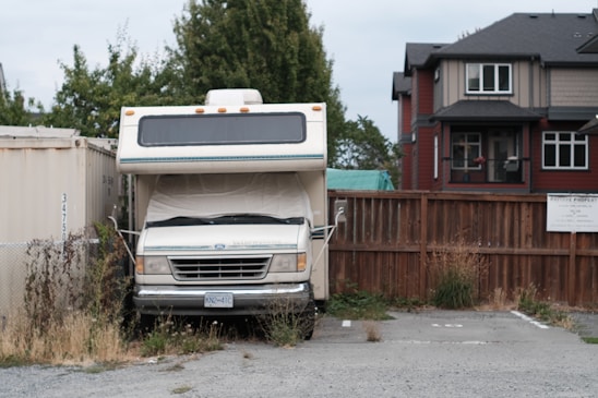 An older model RV is parked in a small, overgrown parking space adjacent to a wooden fence. The RV is flanked by a shipping container and multiple storage units. Weeds and grass are growing around the base of the vehicle. In the background, there is a modern two-story house with dark roofing.