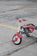 A red and white tricycle with chunky tires is parked on a gray concrete surface. The tricycle appears to be slightly used, with some wear visible on the seat and pedals. Yellow painted lines on the pavement suggest a parking lot or outdoor recreational area.