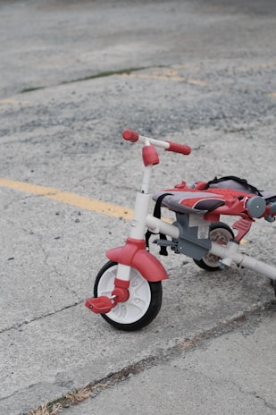 A red and white tricycle with chunky tires is parked on a gray concrete surface. The tricycle appears to be slightly used, with some wear visible on the seat and pedals. Yellow painted lines on the pavement suggest a parking lot or outdoor recreational area.