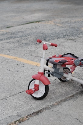 A red and white tricycle with chunky tires is parked on a gray concrete surface. The tricycle appears to be slightly used, with some wear visible on the seat and pedals. Yellow painted lines on the pavement suggest a parking lot or outdoor recreational area.