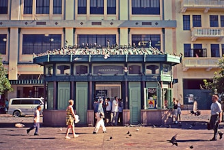 A small, green, kiosk-like building with 'telefonos' written on top is situated in front of a large multi-story building. Several people are walking around or standing near the kiosk, and pigeons are seen both on the roof of the kiosk and on the ground. The background building has multiple windows and a yellow hue. A van is parked to the left of the kiosk.