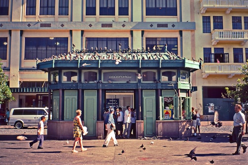 A small, green, kiosk-like building with 'telefonos' written on top is situated in front of a large multi-story building. Several people are walking around or standing near the kiosk, and pigeons are seen both on the roof of the kiosk and on the ground. The background building has multiple windows and a yellow hue. A van is parked to the left of the kiosk.