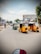 Three auto-rickshaws are driving on a busy urban street with shops, signs, and trees in the background. There is a green auto-rickshaw along with two yellow ones. Other vehicles like a motorbike and a car are also visible on the road, and wires hang overhead across the street.