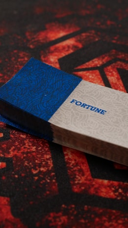 A stack of cards is placed on a surface featuring a red and black geometric pattern. The top card has a blue and white design with the word 'FORTUNE' written in blue. The card stack appears slightly worn with textured edges.