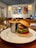 A plated meal featuring a burger with a variety of toppings next to a metal cup filled with French fries. The setting appears to be a cozy dining area with wooden tables and artistic framed prints on the walls, illuminated by hanging lights. A glass pitcher is placed nearby, containing a drink with mint leaves and lemon slices.